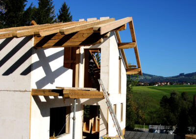 Blick auf einen Neubau mit montiertem Altholz-Dachstuhl, von rechts fotografiert mit grüner Wiese, Baustellen-Elementen und blauem Himmel im sonnigen Hintergrund.