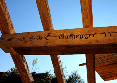 Von unten in den blauen Himmel fotografiert, zeigt das Bild vier massive handgehackte Altholz-Sparren und eine mit „Buchegger“ gravierte Pfette auf einer Baustelle.