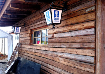 Außenansicht einer Altholz-Fassade mit Sparren in Blockbau-Optik mit zwei rustikalen Augustiner Bräu München Lampen neben einem noch unverkleideten Fenster.