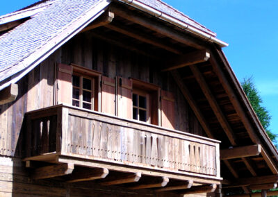 Detailaufnahme einer fertigen Altholzfassade aus gehackten Brettern mit einem passenden Holzbalkon vor strahlend blauem Himmel.