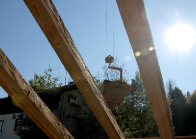 Detailaufnahme von unten in den blauen Himmel fotografiert, zeigt drei massive, handgehackte Altholz-Sparren, die von einem Kranhaken in Position gebracht werden.