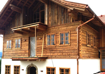 Außenansicht eines Einfamilienhauses mit gemauertem Erdgeschoss, Obergeschoss in Blockbau-Optik und einer Fassade aus gehackten Altholzbrettern im Dachgeschoss.