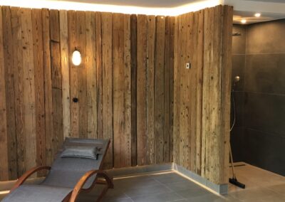 Wellnessbereich mit handgehackter Altholzwand, indirekter LED-Beleuchtung, Wellnessliege und modern gefliester Dusche.