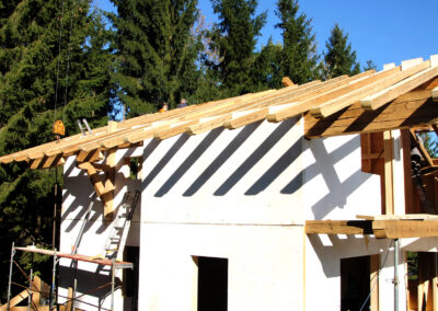 Neubau mit Altholz-Dachstuhl: Tradition trifft Moderne Alternativer Text: Seitliche Ansicht eines Neubaus in der Bauphase, dessen Dachstuhl mit massiven, handgehackten Altholz-Sparren errichtet wird, mit Kran und Fichtenwald im Hintergrund unter blauem Himmel.