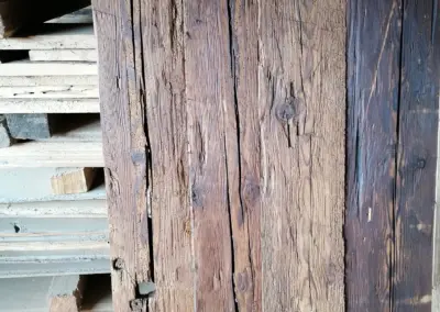 Muster-Präsentation von vier hochgestellten, dunkel sortierten Altholzbrettern mit gehackter Oberfläche neben einem Holzstapel.